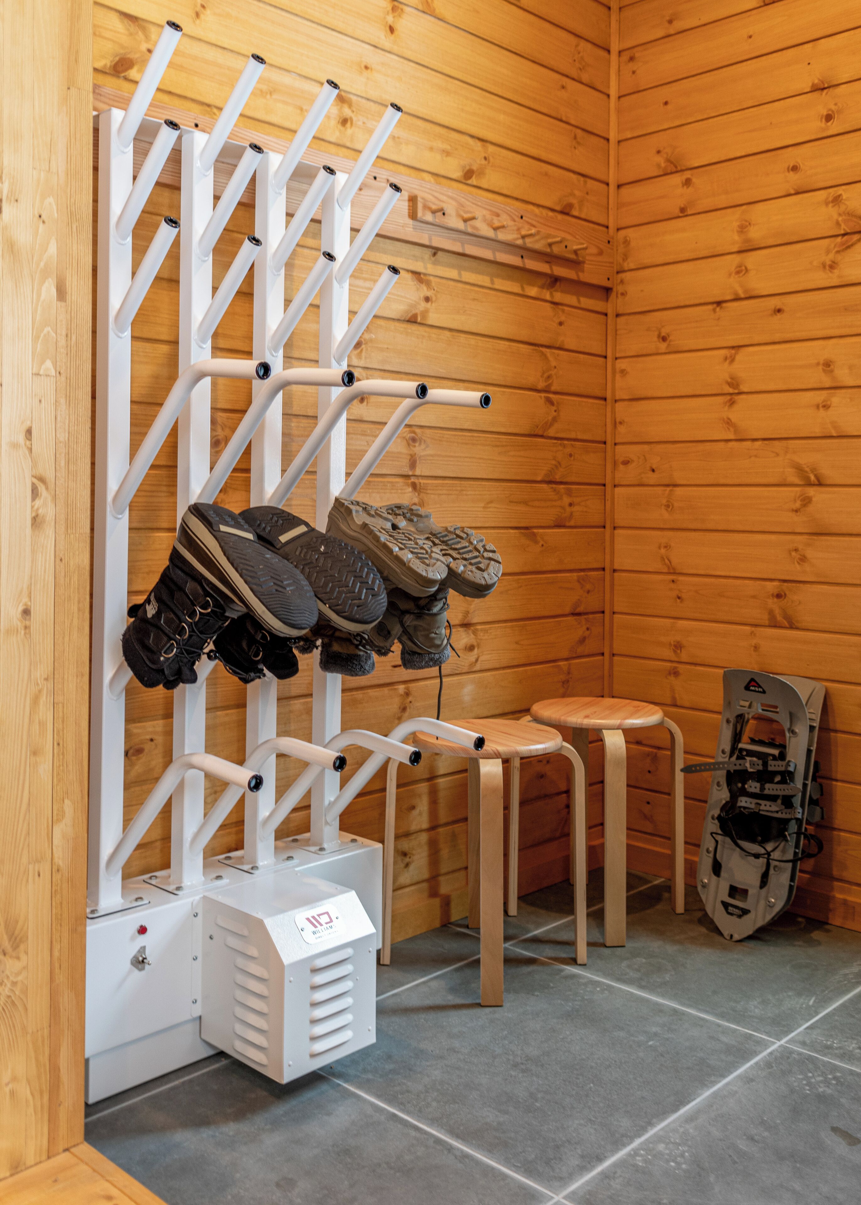 A wooden room with a shoe rack holding boots, a small stool, and a snowboard leaning against the wall.