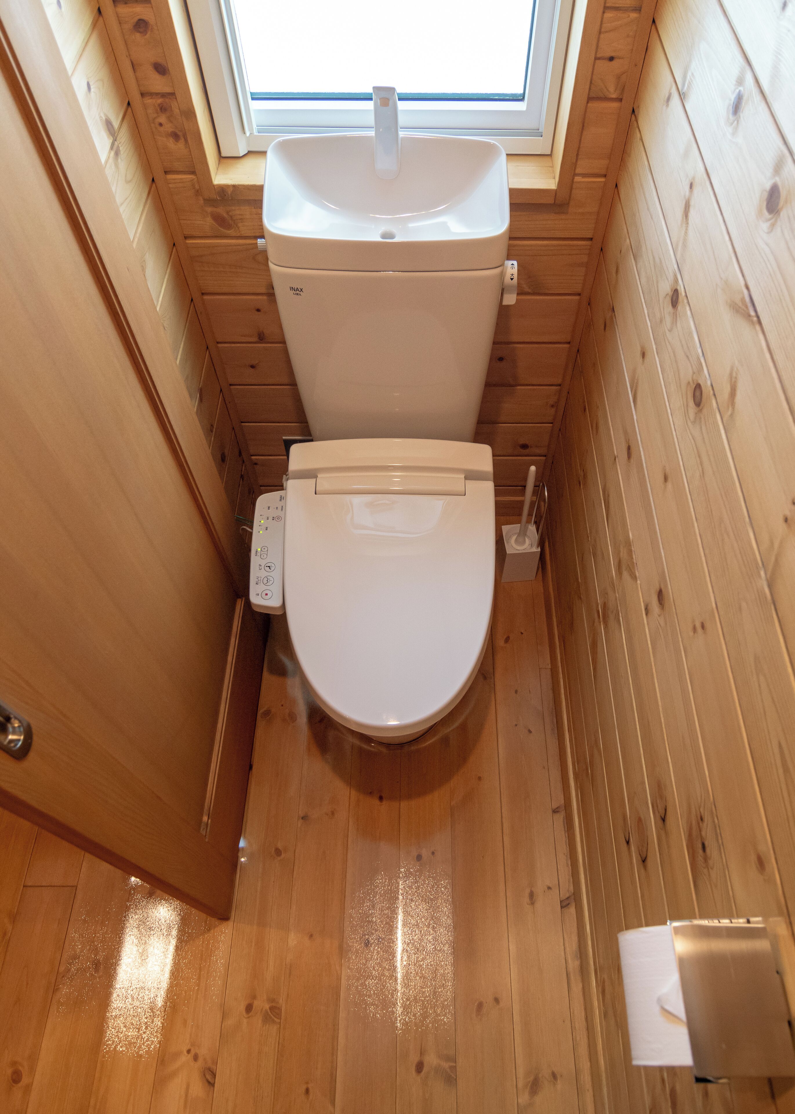 Une petite salle de bain aux murs lambrissés, avec des toilettes blanches et une fenêtre située juste au-dessus.