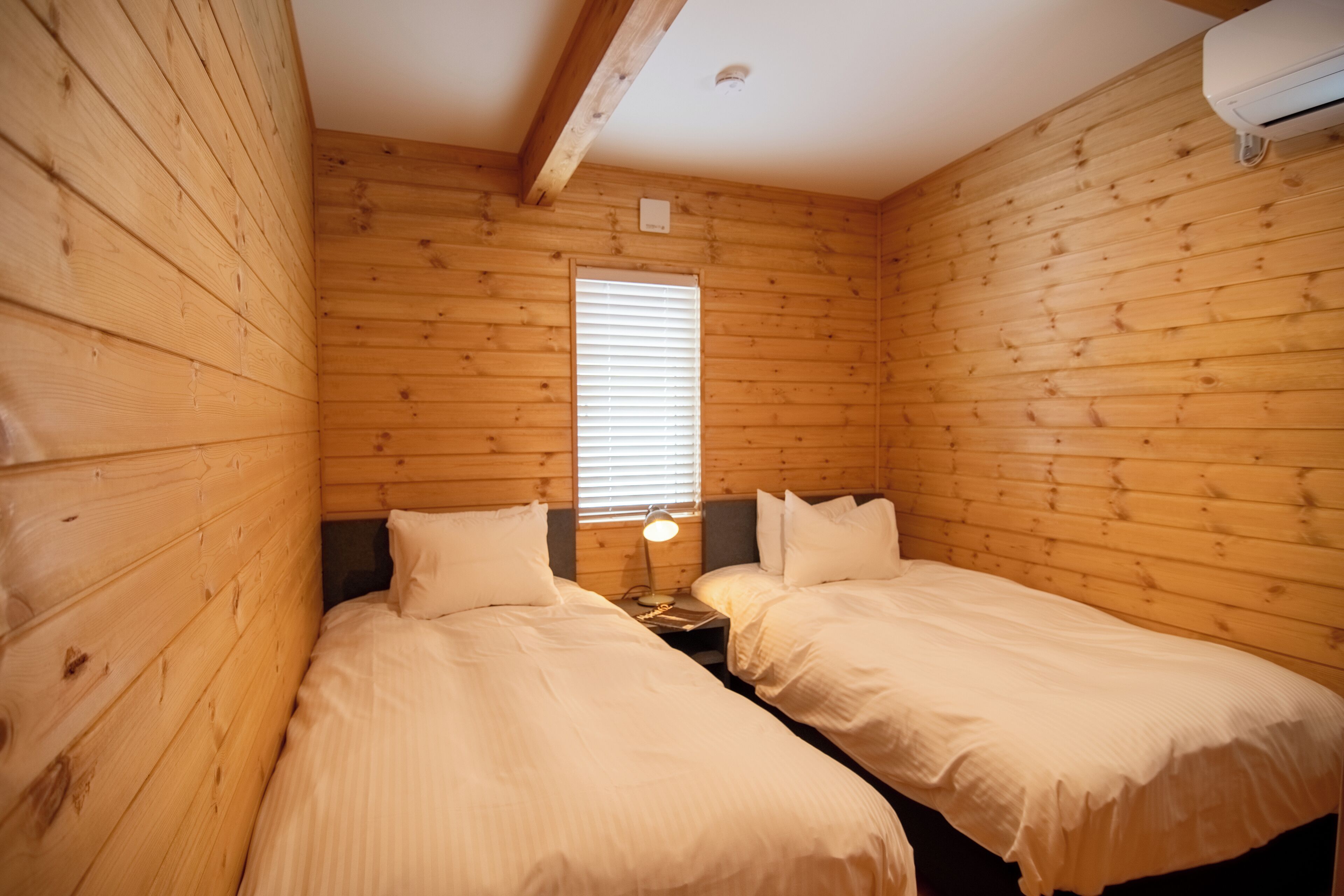 A wooden cabin room with two beds, a window, and a nightstand with a lamp.