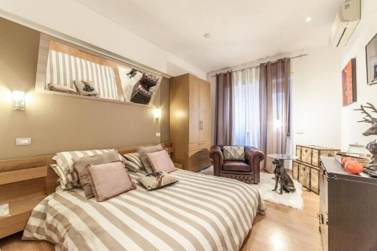 A bedroom with a large bed, a wooden wardrobe, a leather armchair, a small table with a dog statue, and a desk with a red object.