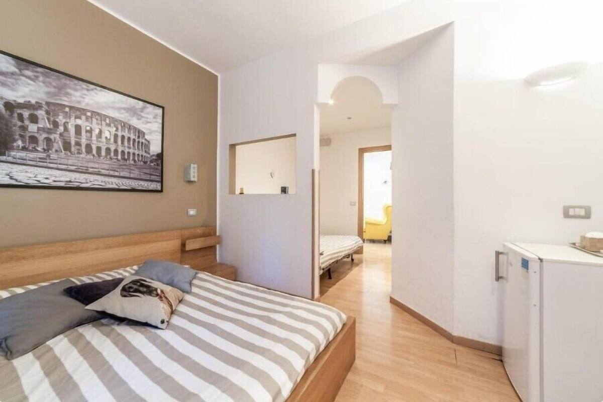 A bedroom with a bed, a small desk, a refrigerator, and a picture of the Colosseum on the wall.