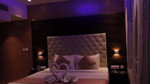 A hotel room with a tufted headboard, a bed with a heart-shaped towel arrangement, and two wall-mounted lamps. - NPG Hotel (KA)
