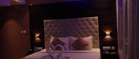 A hotel room with a tufted headboard, a bed with a heart-shaped towel arrangement, and two wall-mounted lamps.