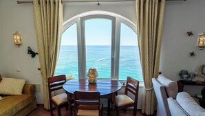A room with a large arched window overlooking the sea, a round wooden table with chairs, a sofa, and a decorative lamp.