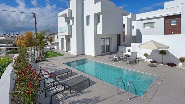 A modern pool area with a clear blue pool, lounge chairs, and a white building with a flat roof. - Sun & Moon Luxury Villas Ayia Napa (Ayia Napa)