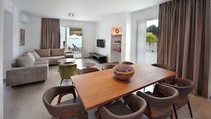 A modern living room with a sofa, dining table, and chairs. There is a TV on the wall, a window with curtains, and a balcony with outdoor furniture. - Sun & Moon Luxury Villas Ayia Napa (Ayia Napa)
