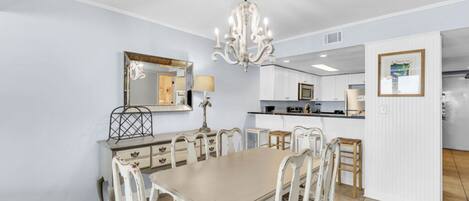 A dining area with a chandelier, a mirror, and a kitchen in the background.