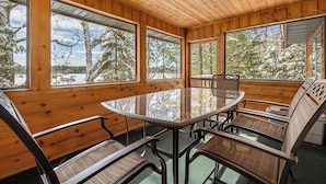 Ein Zimmer mit Holzwänden, einem Glastisch und Stühlen, großen Fenstern und Blick auf schneebedeckte Bäume.