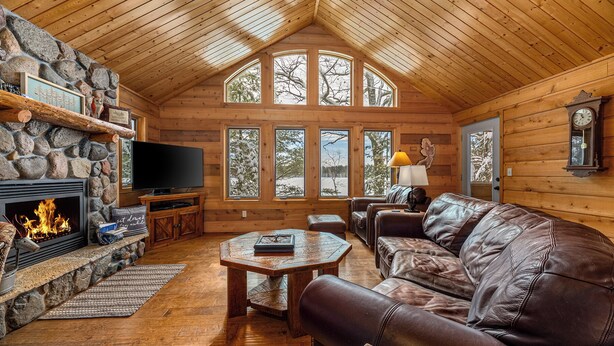 Ein gemütliches Blockhaus-Interieur mit Kamin aus Stein, Ledersofa und großen Fenstern mit Blick auf schneebedeckte Bäume.