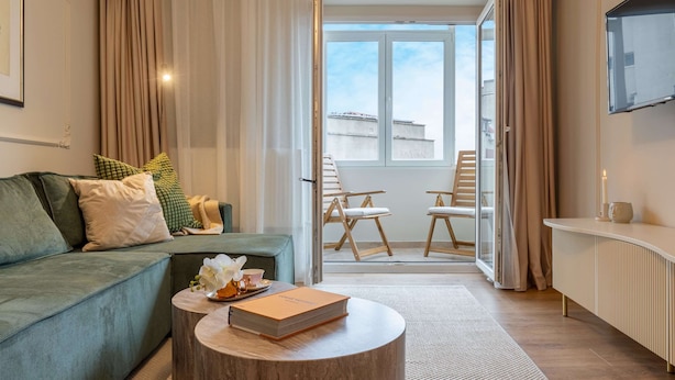 A modern living room with a green sofa, a round wooden coffee table, and a view of a balcony with outdoor seating.