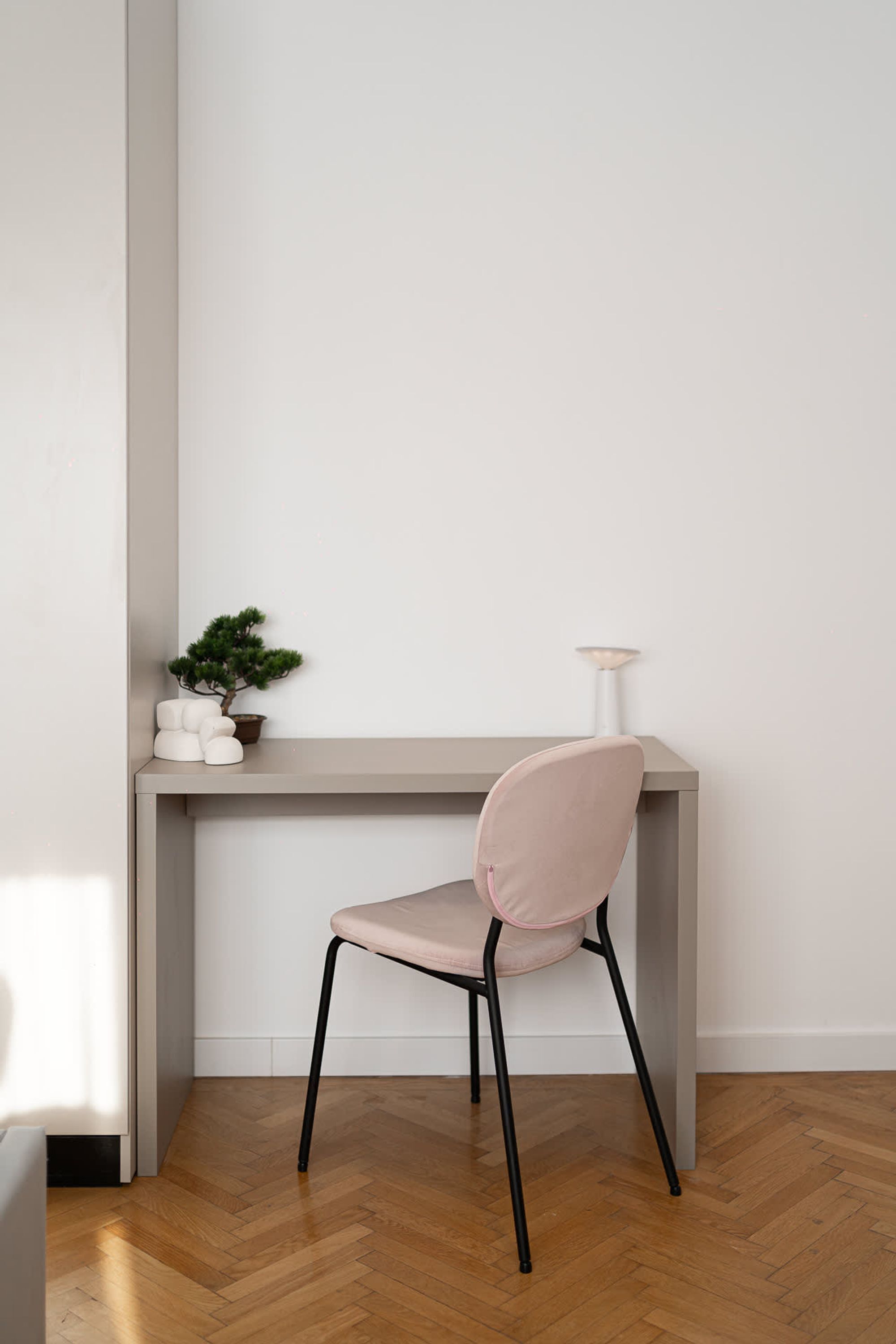A minimalist room with a wooden herringbone floor, a grey desk with a small potted plant, a pink chair, and a white lamp.