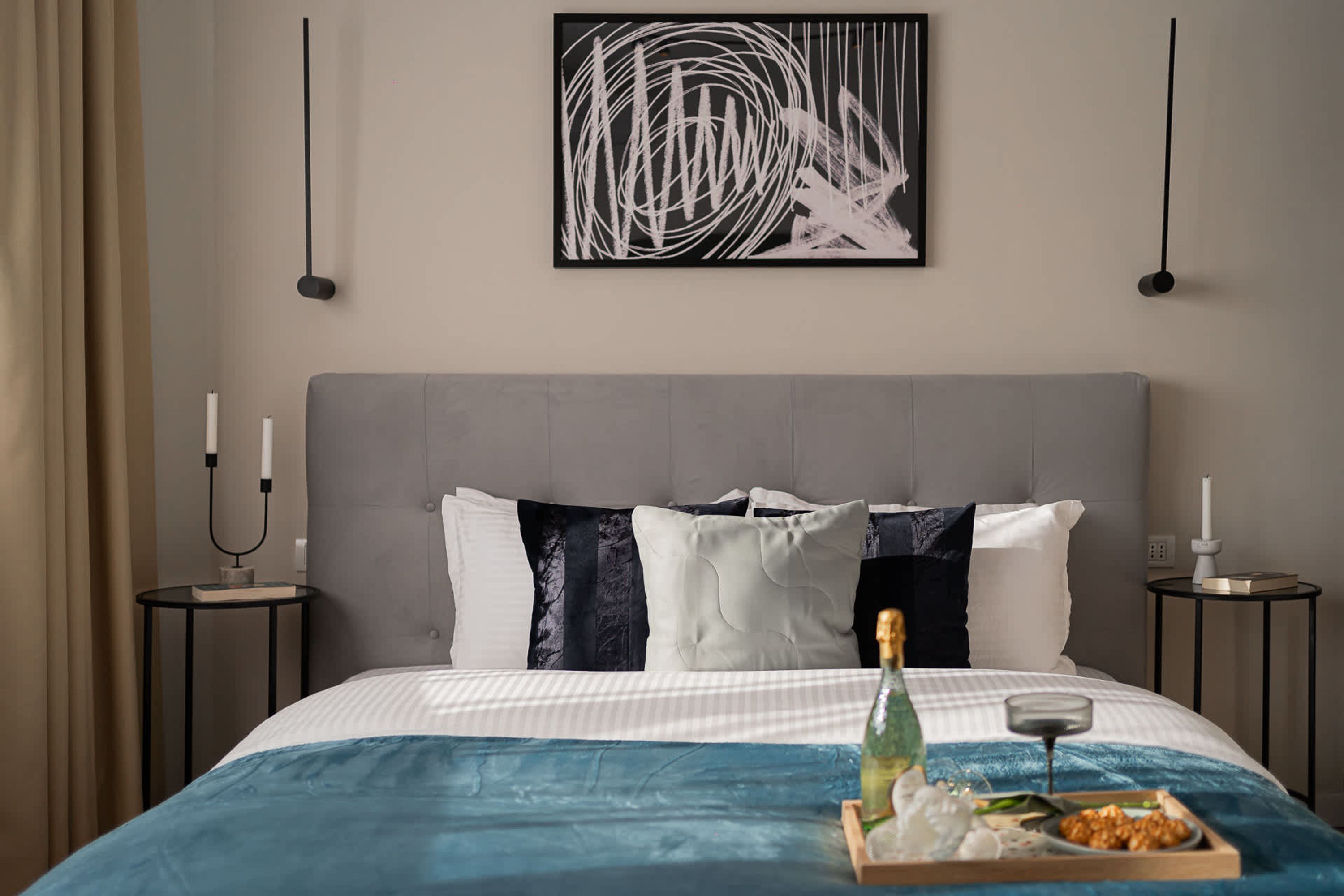 A neatly made bed with a blue bedspread, white pillows, and a bottle of champagne on the side.