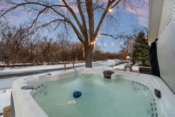 A hot tub with a blue floating device, surrounded by snow, trees, and string lights. - Cozy 3BR Cabin With Hot Tub on Nippersink Creek (Spring Grove)