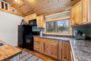 A kitchen with wooden cabinets, a black refrigerator, a stove, and a window with a view of trees. - Cozy 3BR Cabin With Hot Tub on Nippersink Creek (Spring Grove)