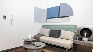 A modern hotel room with a sofa, a round coffee table, a side table, a wall-mounted light fixture, and a wall-mounted thermostat.