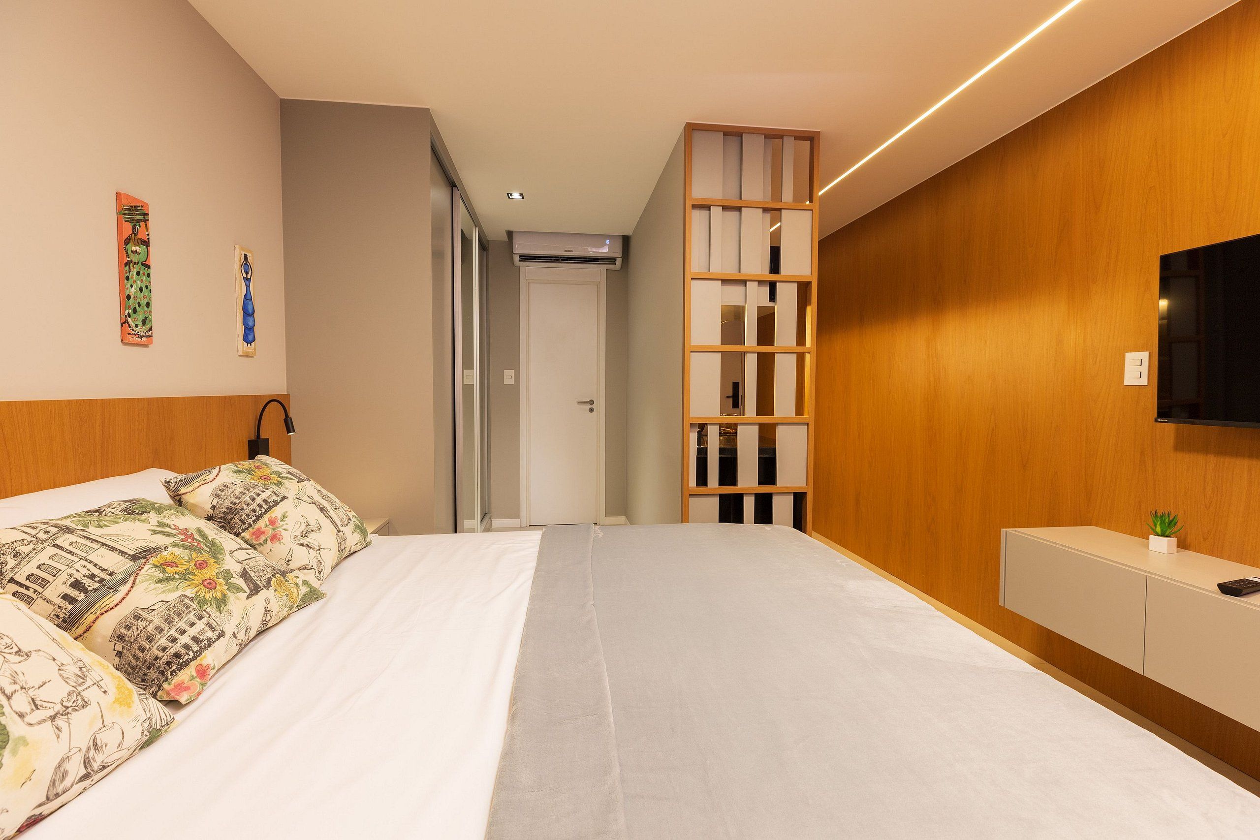 A modern hotel room with a large bed, a wooden panel wall, a mounted TV, and a small plant on the nightstand.