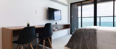Una habitación de hotel moderna con una cama grande, un escritorio con dos sillas, un televisor de pantalla plana montado en la pared y vistas al mar a través de ventanas amplias.