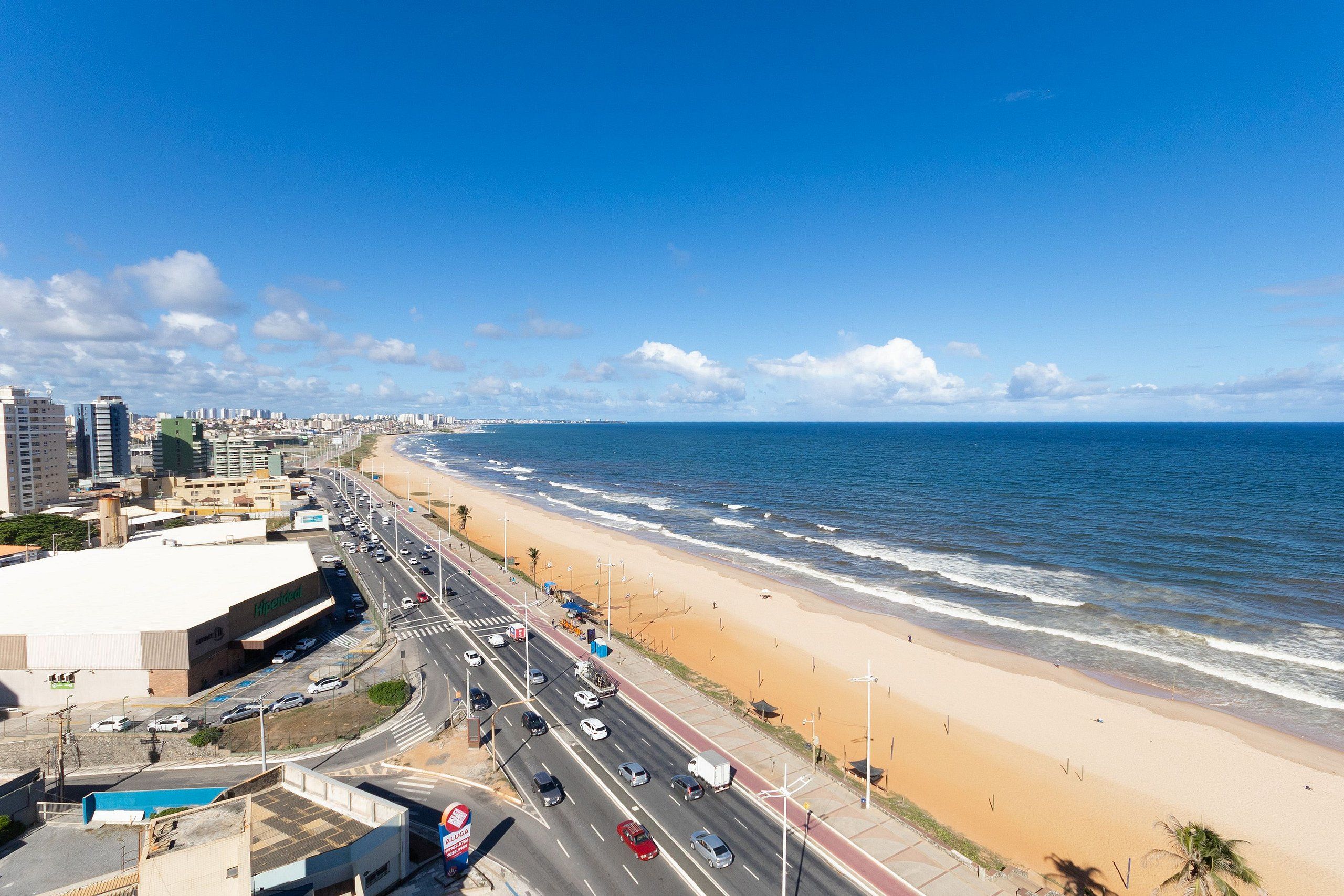 Un paisaje urbano costero con playa, una carretera con vehículos y un edificio grande con un letrero.