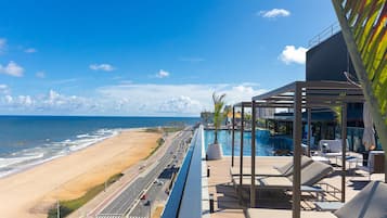 A balcony with a view of the beach and ocean, lounge chairs, and a pool area.