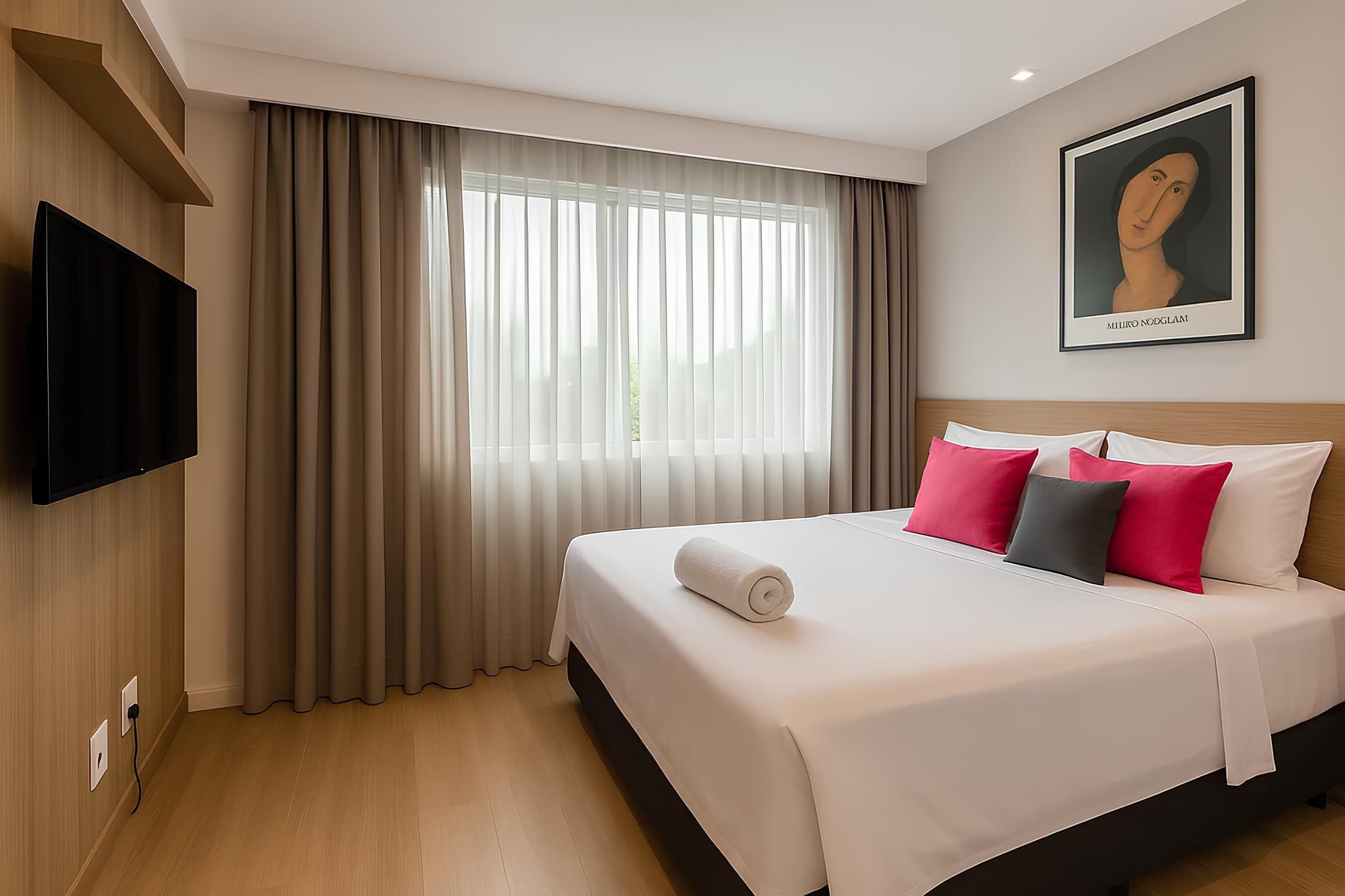 A hotel room with a large bed, a flat-screen TV mounted on the wall, and a window with sheer curtains.