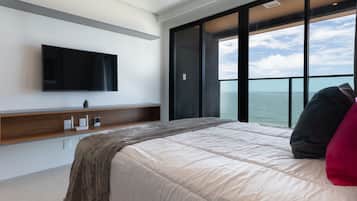 A modern bedroom with a large bed, a flat-screen TV mounted on the wall, a wooden console table, and a view of the sea through large windows.