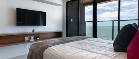 A modern bedroom with a large bed, a flat-screen TV mounted on the wall, a wooden console table, and a view of the sea through large windows.
