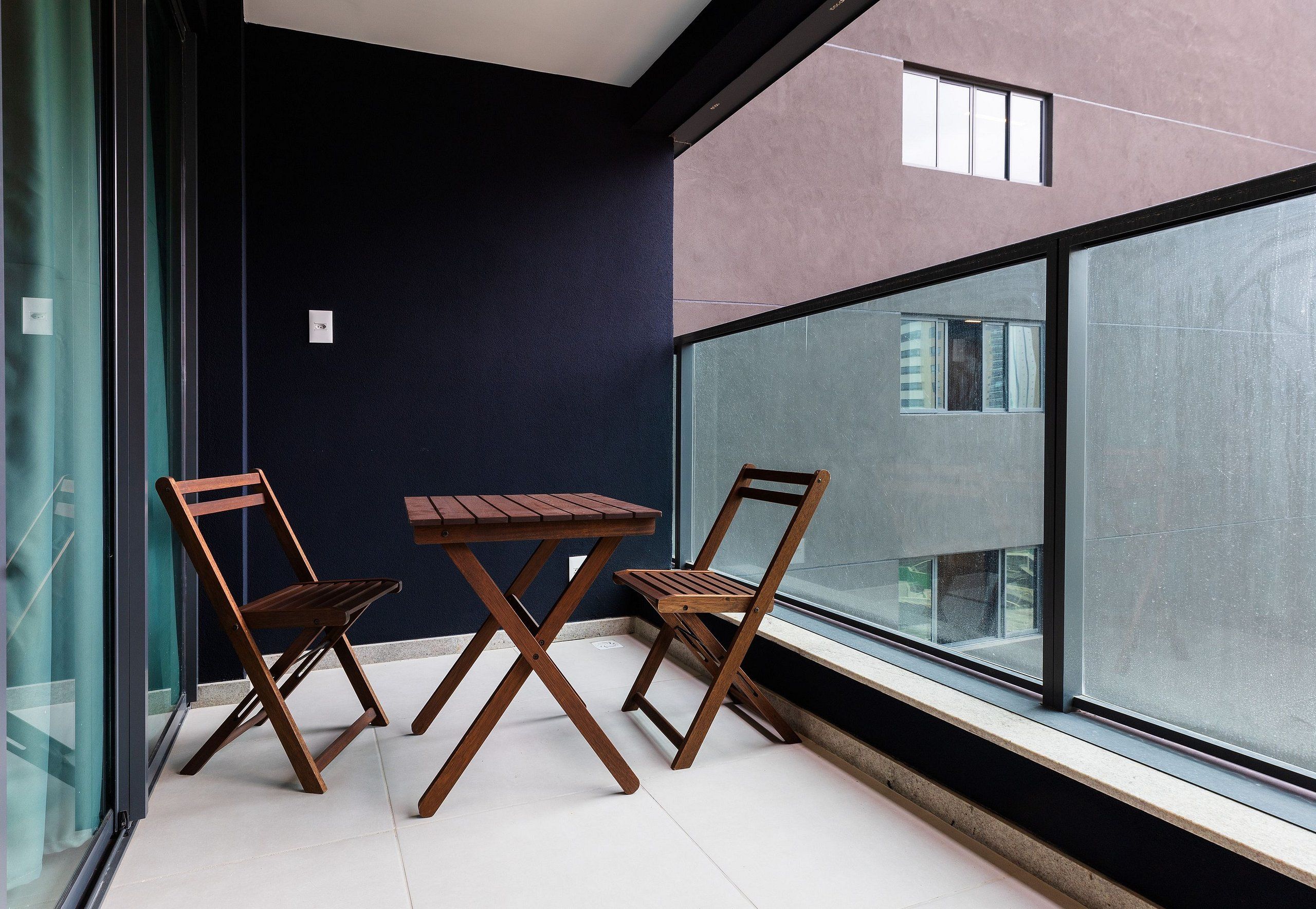 Un balcone con due sedie di legno e un tavolo, una porta in vetro e vista su un edificio attraverso ampie finestre.