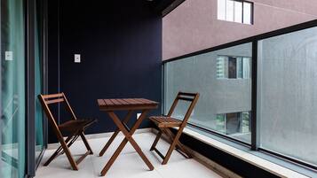 Un balcone con due sedie di legno e un tavolo, una porta in vetro e vista su un edificio attraverso ampie finestre.