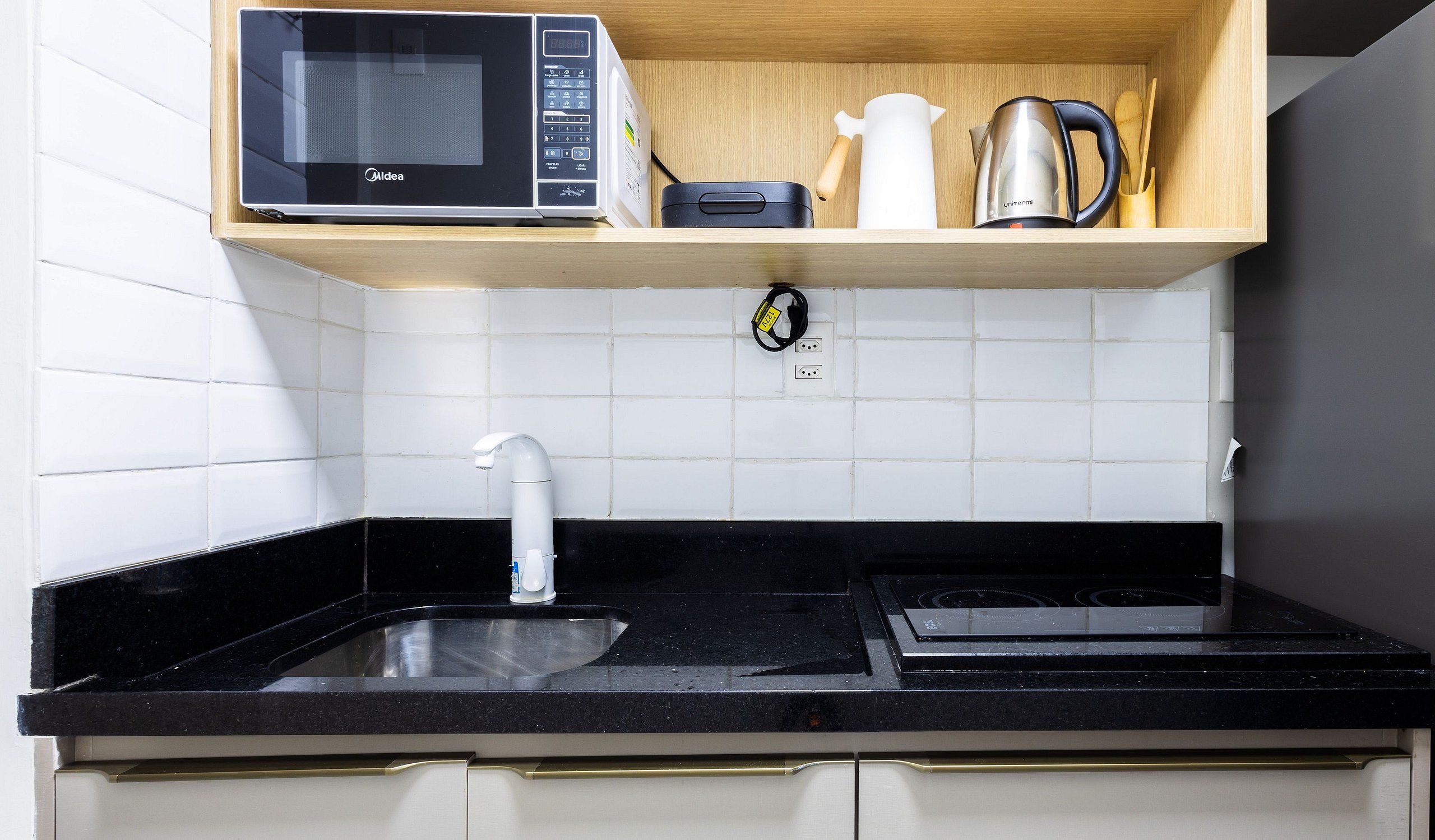 A kitchen with a black countertop, a stainless steel sink, a microwave, and a kettle on a wooden shelf.