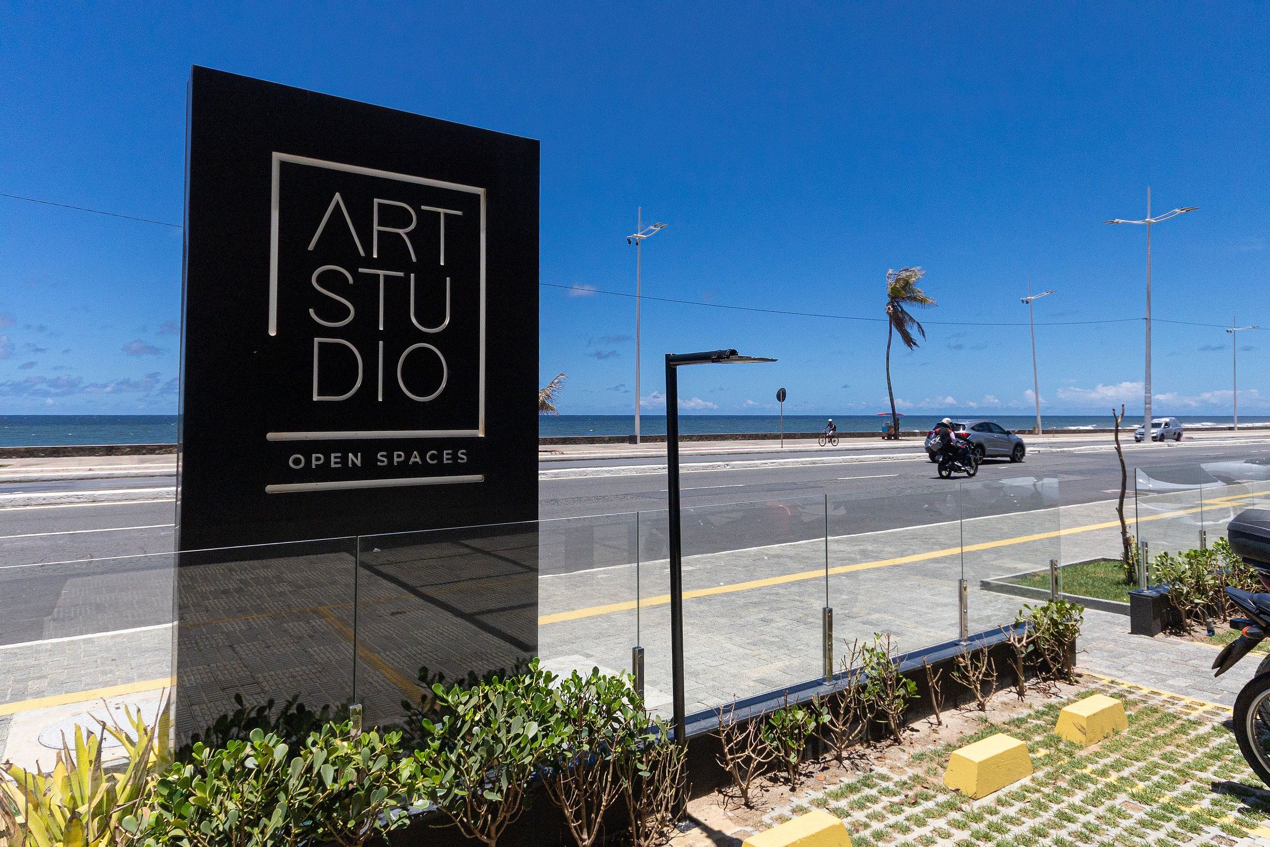 Een bord met 'ART STUDIO' erop, en daaronder 'OPEN SPACES', gelegen nabij een weg met vrij uitzicht op de oceaan en palmbomen op de achtergrond.