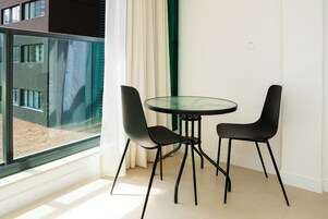 Quarto moderno com mesa de vidro e duas cadeiras, uma janela grande com cortinas e vista para um edifício lá fora.