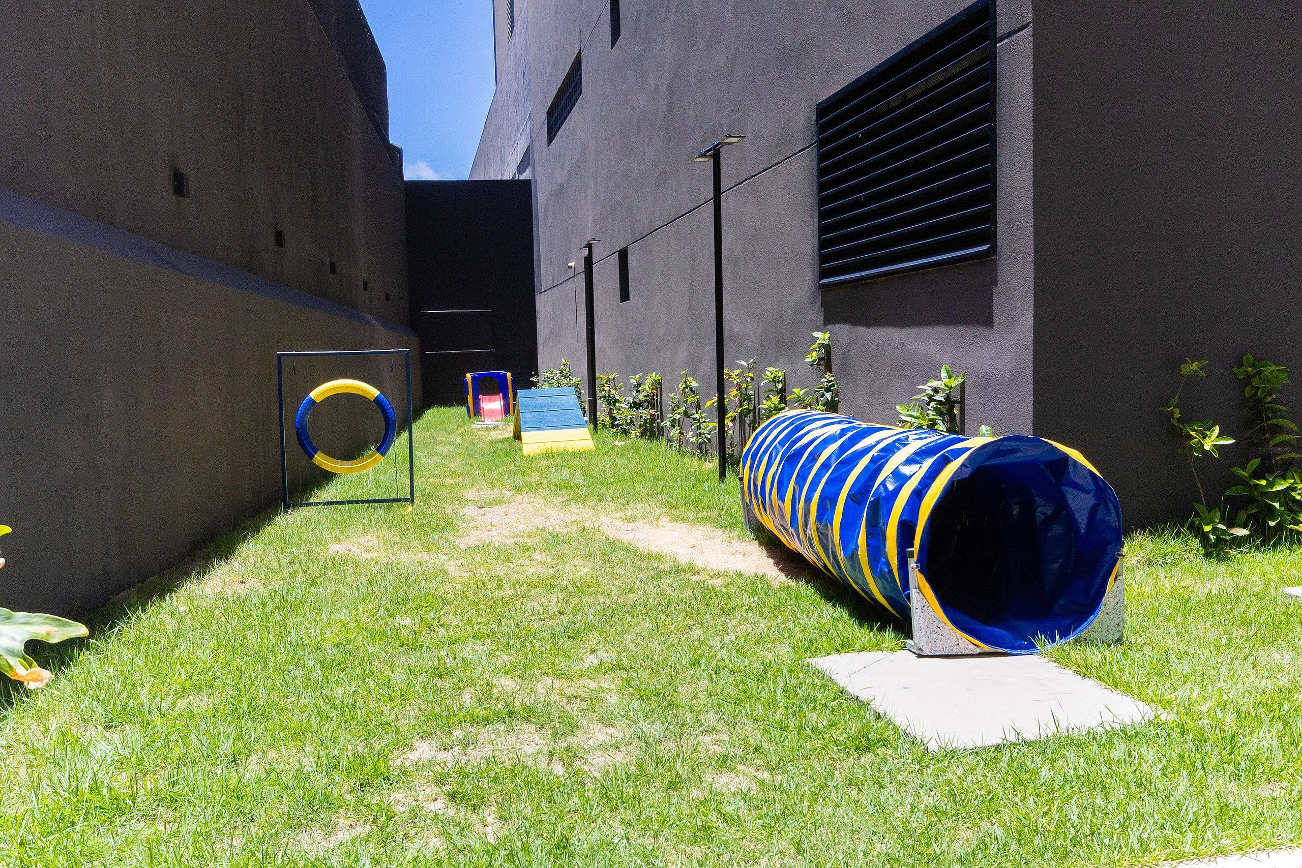 Uma área externa gramada com um túnel azul e amarelo, um escorrega amarelo e uma estrutura de escalada azul e amarela.