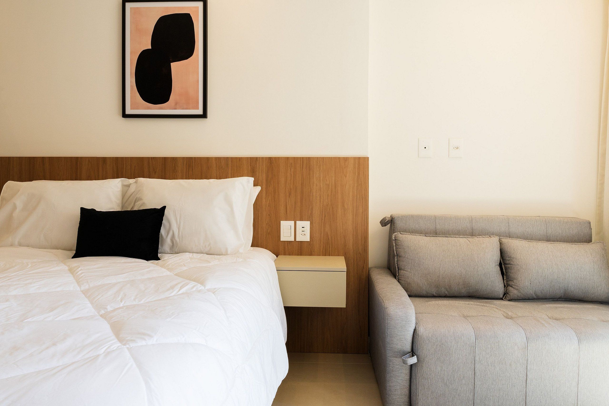 A hotel room with a bed, a bedside table, a gray sofa, and a framed abstract artwork on the wall.