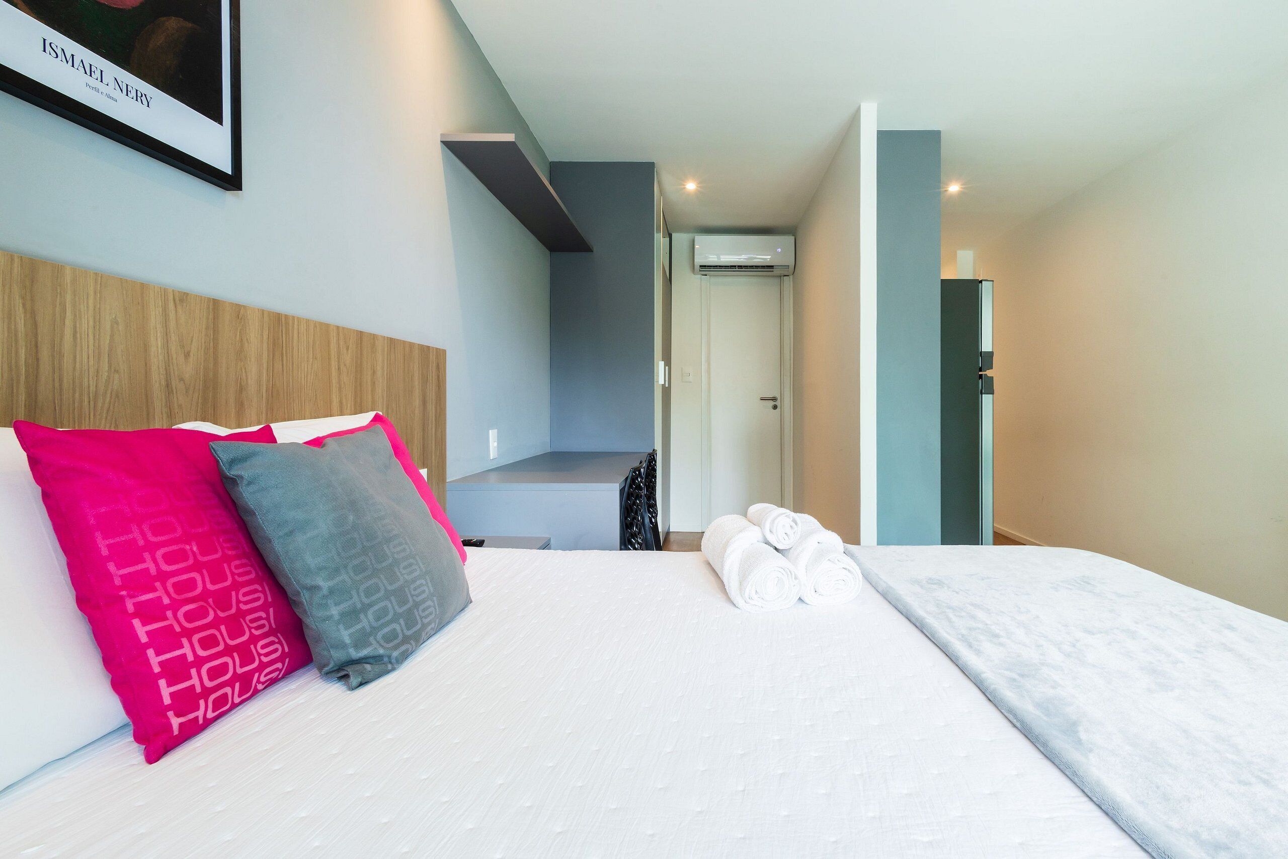 A hotel room with a bed, pillows, a framed picture, and a door leading to another room.