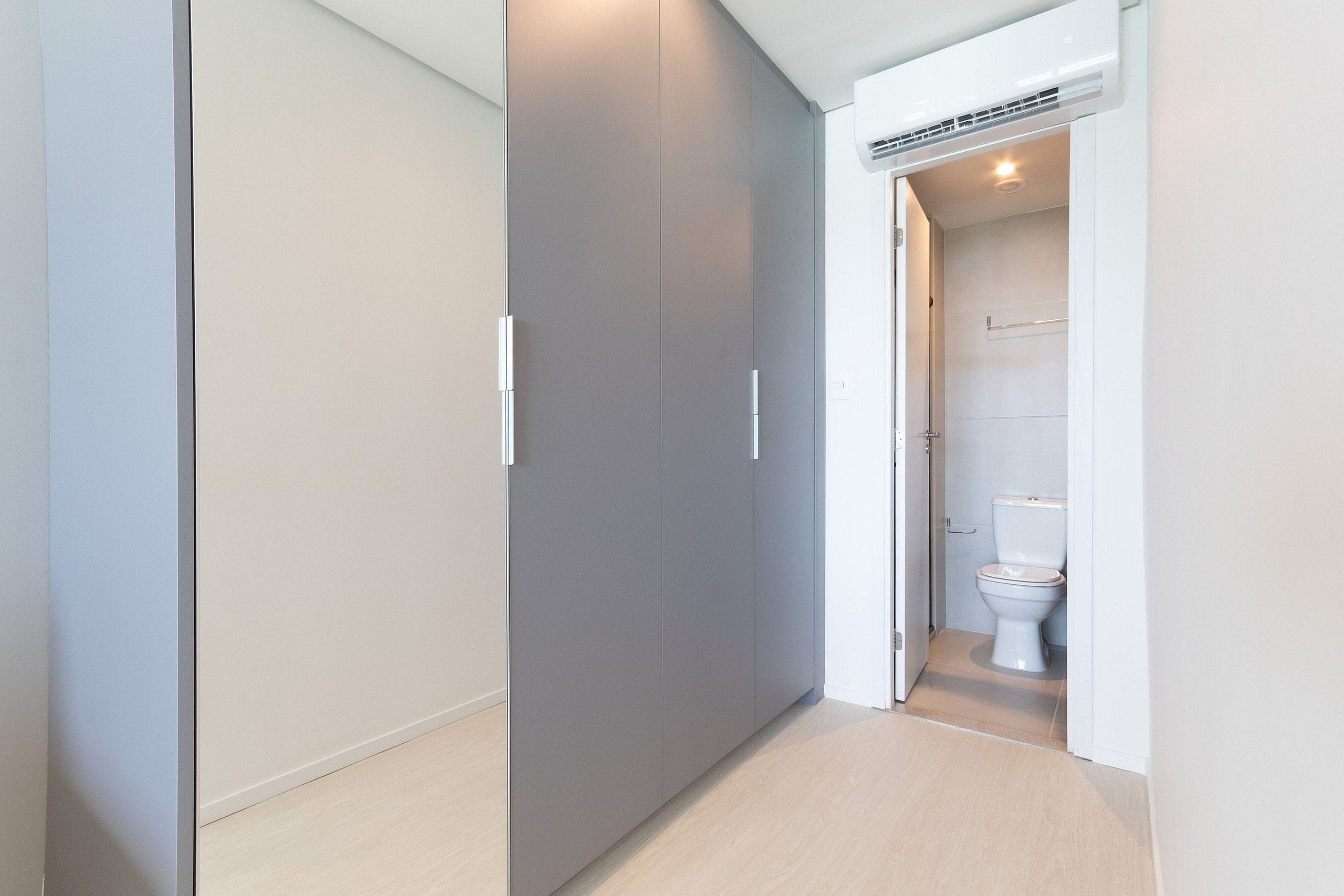 Um corredor com um guarda-roupa cinzento, uma porta branca que dá para um banheiro com vaso sanitário e chuveiro.