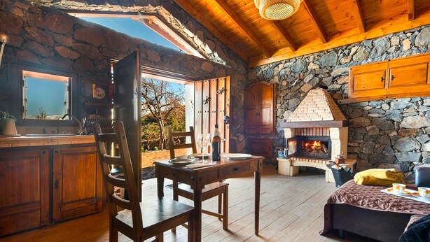 A rustic cabin interior with a stone fireplace, wooden furniture, and a large window overlooking a garden. - Holiday Home in Tenerife With Private Garden (Santa Ursula)