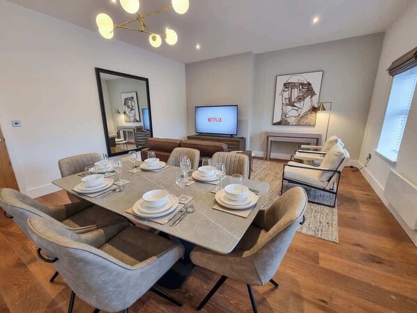 A modern dining area with a large table set for six, a sofa, and a TV displaying 'NETFLIX'.