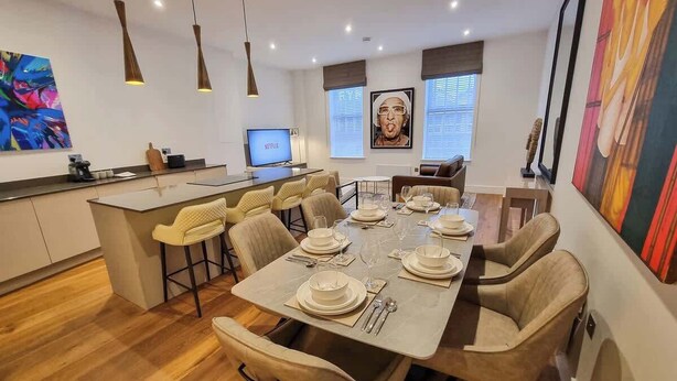 Un espace de salle à manger moderne avec une longue table dressée pour six personnes, une cuisine en arrière-plan et des œuvres d’art accrochées aux murs.