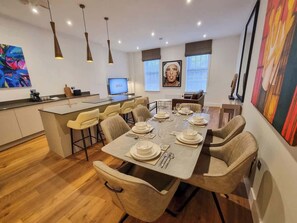 A modern dining area with a long table set for six, a kitchen in the background, and artwork on the walls. - 2 Bed Altrincham Centre | 2 Bath | Sleeps 6 (Greater Manchester)