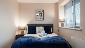 A bedroom with a bed, pillows, a bedside lamp, a wall art piece, and a window with blinds. - 1 Bed Media City Flat | Free Parking (Manchester)