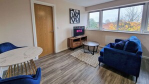 A living room with a TV, a wooden door, a window with a view, a blue sofa, and a small table. - 1 Bed Media City Flat | Free Parking (Manchester)