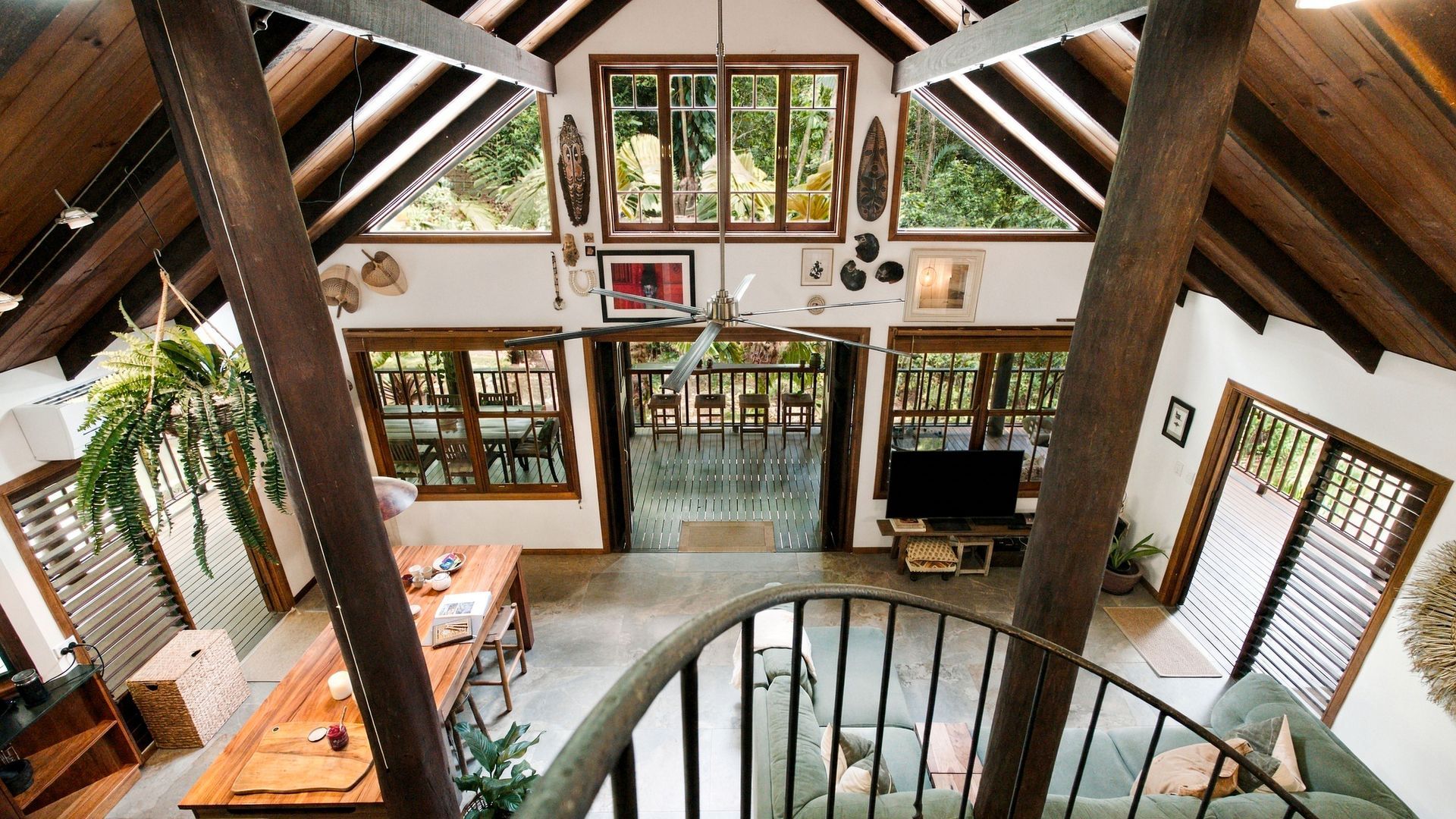 A spacious room with a high ceiling, wooden beams, a central staircase, a dining area with a table and chairs, and large windows offering views of the outdoors.
