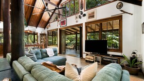 A spacious living area with a high ceiling, a large flat-screen TV, and a view of lush greenery through large windows. - Fern House your serene rainforest retreat (Bingil Bay)