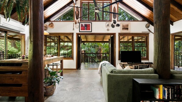 A spacious living area with a large window, a television, and a bookshelf. - Fern House your serene rainforest retreat (Bingil Bay)