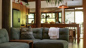 A cozy living room with a green sofa, wooden furniture, and a kitchen area with a window.