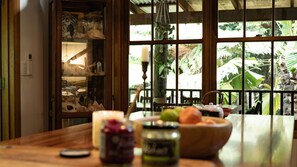A dining area with a wooden table, a bowl of fruit, and a view of a garden through large windows. - Fern House your serene rainforest retreat (Bingil Bay)