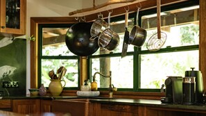 A kitchen with hanging pots, a window with a view of greenery, and various kitchen utensils on the counter.