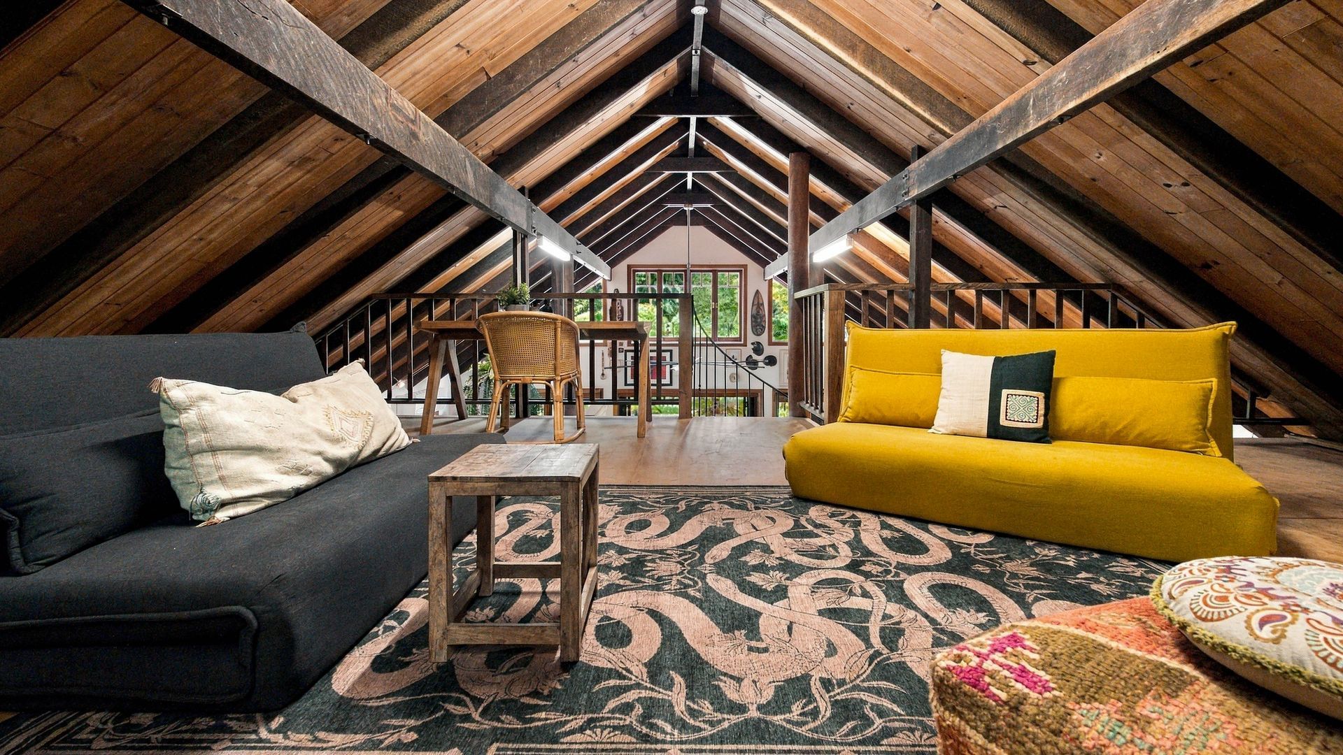 An attic room with a high ceiling, wooden beams, a black sofa, a yellow sofa, a wooden coffee table, and a patterned rug.