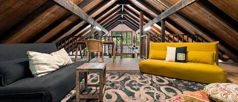 An attic room with a high ceiling, wooden beams, a black sofa, a yellow sofa, a wooden coffee table, and a patterned rug.