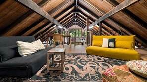 An attic room with a high ceiling, wooden beams, a black sofa, a yellow sofa, a wooden coffee table, and a patterned rug.